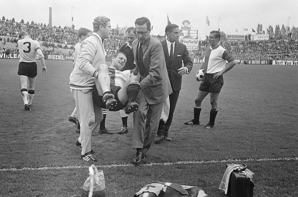 Feyenoord speler Piet Fransen wordt buiten de lijnen gedragen, rechts trainer Willy Kment. Foto: fotograaf onbekend / Anefo, Nationaal Archief (CC0).